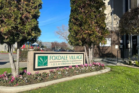 A sign for Foxdale Village with a phone number and address is surrounded by trees and flowers.