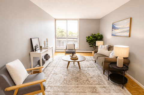A living room with a grey rug, a grey sofa, a wooden chair, a wooden coffee table, a white side table, a white wall, a window, a plant, and a painting.