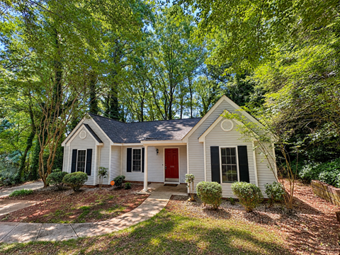 A small white house with a red door is surrounded by green trees.