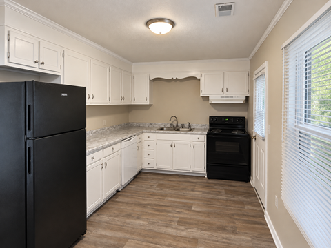 A black refrigerator stands in a kitchen with white cabinets and a wooden floor.
