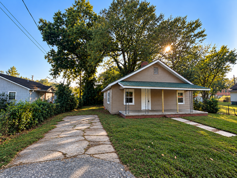 A house with a green roof is surrounded by trees and has a driveway leading to the front door.