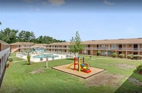 A playground with a slide and a tree in the middle of a grassy area in front of a building.