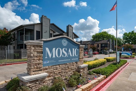 The image shows a sign for MOSAIC in front of a building with a flag on a pole.