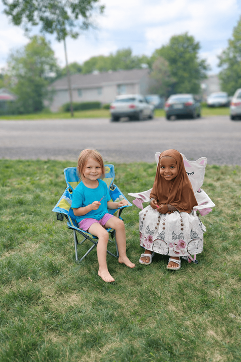 Two children sitting on chairs on the grass.