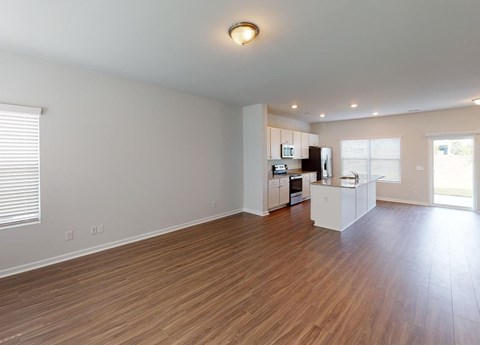 A spacious living room with wooden flooring and a kitchen area in the background.