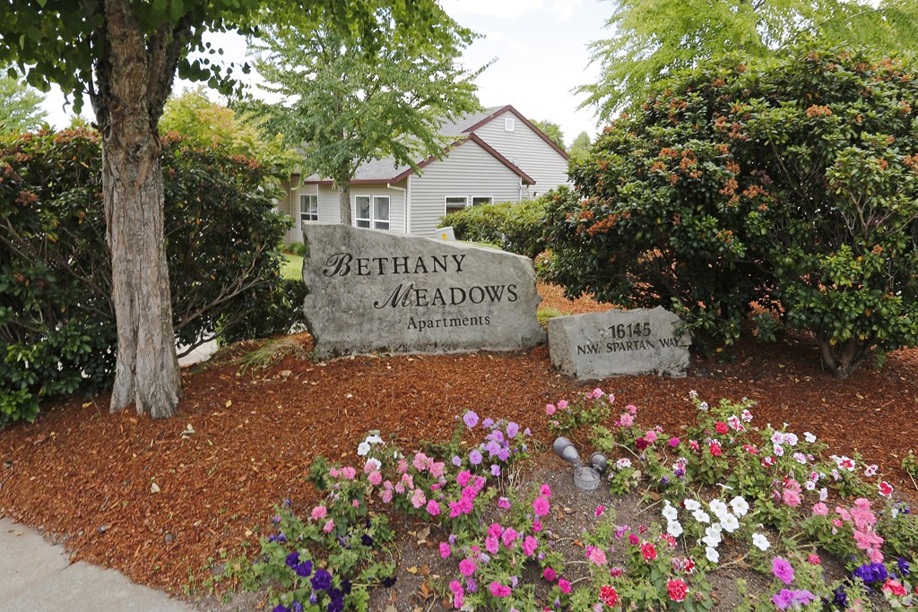 A sign for Bethany Meadows Apartments is surrounded by flowers and trees.