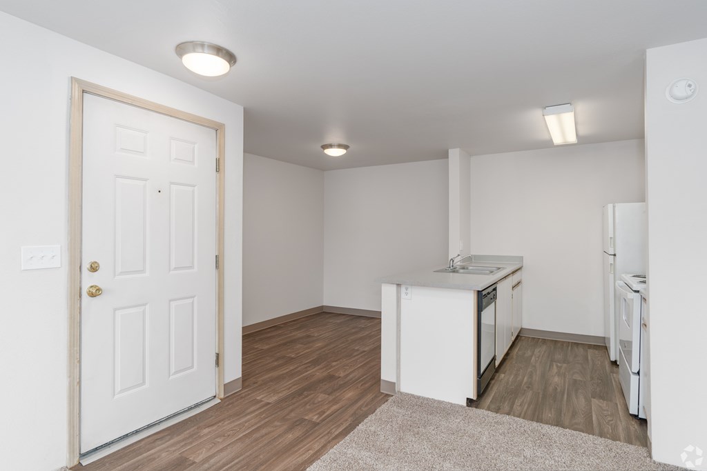 A white door is on the left of a room with a kitchenette.