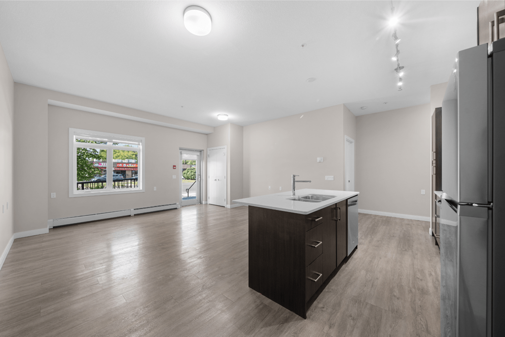 an empty living room with a kitchen with a refrigerator and a sink