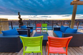 A balcony with a view of the city at dusk, furnished with blue cushions and colorful chairs.