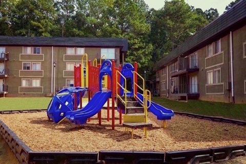 A playground with a blue slide and yellow and red play structure in front of apartment buildings.