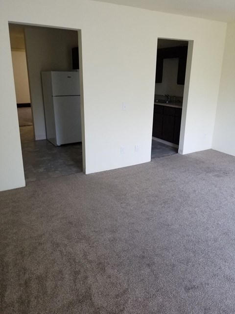 a living room with carpet and a kitchen with a refrigerator