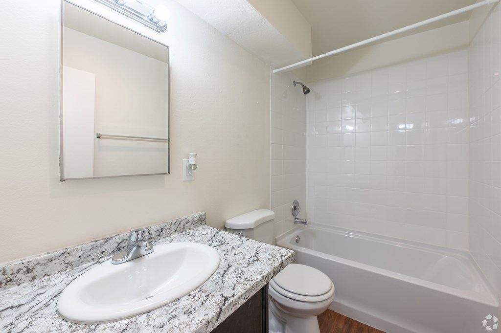 A bathroom with a marble countertop, a white toilet, a sink, a mirror, and a tub with a shower.