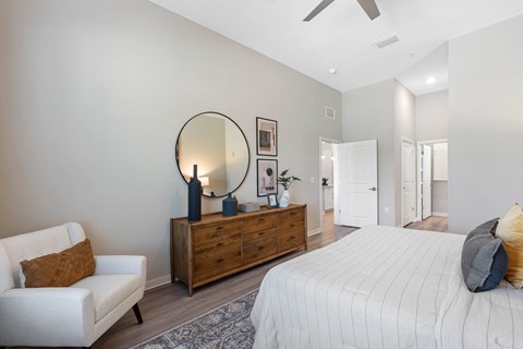 A bedroom with a large bed, a white chair, and a wooden dresser with a mirror above it.