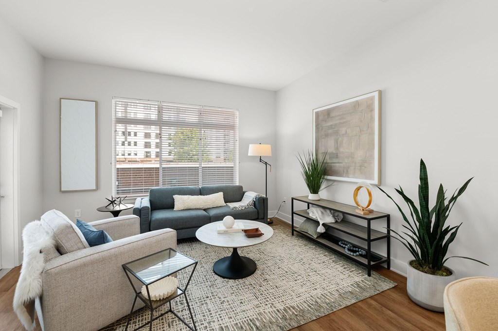 A living room with a grey couch, a glass table, a white chair, a green plant, and a painting on the wall.