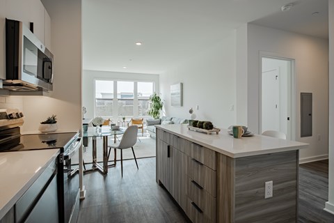 Open concept kitchen and living room at The Rail Bound Brook with wood-style flooring, stainless steel appliances, and large sunlit windows.