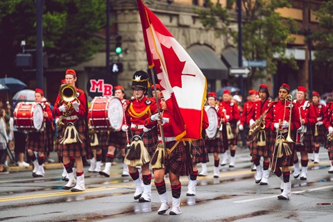 A group of men in red and black kilts marching down a street holding a Canadian flag.