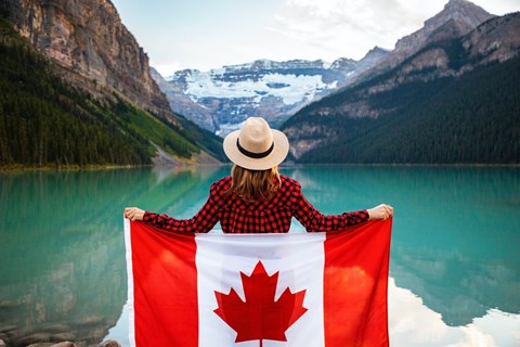 A woman in a hat is holding a Canadian flag in front of a lake and mountains.
