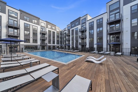 A large wooden deck with sun loungers and a pool in front of apartment buildings.