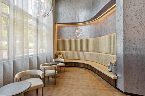 A curved bench with a wooden back and a cushion sits in a room with a grey wall and a window with white curtains.