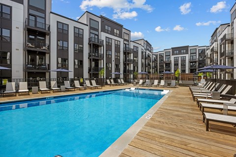 A large swimming pool surrounded by lounge chairs and umbrellas in front of apartment buildings.