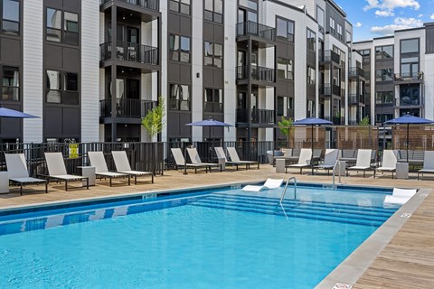 A pool surrounded by chairs and umbrellas in front of apartment buildings.