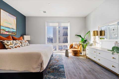 A bedroom with a large bed, a chair, a dresser, and a window with a view of a city skyline.