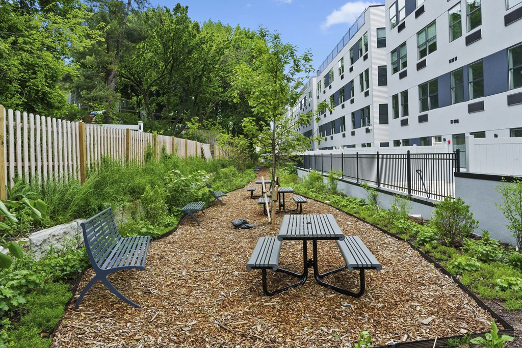 A gravel patio with picnic tables and chairs is surrounded by a fence and trees.