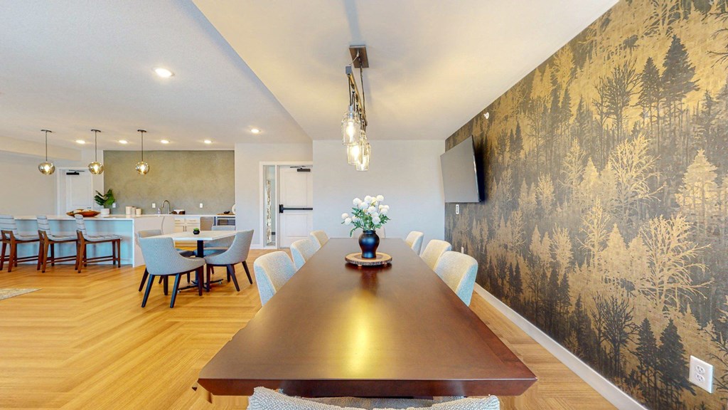 A long wooden table is surrounded by chairs in a room with a forest wallpaper.