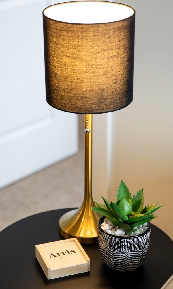 A table lamp with a gold base and a brown lampshade sits on a table with a small potted plant and a book titled