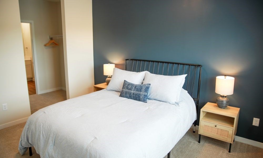 A bed with a white comforter and a blue pillow is in a room with a lamp on a nightstand.