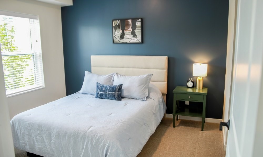 A bed with a white comforter and two blue pillows sits in a room with a green nightstand and a framed picture on the wall.