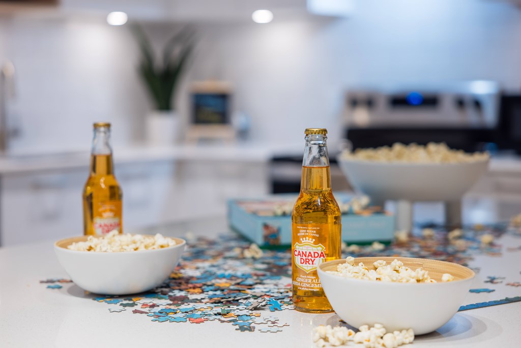 Two bottles of beer are on a table with bowls of popcorn and puzzle pieces scattered around.