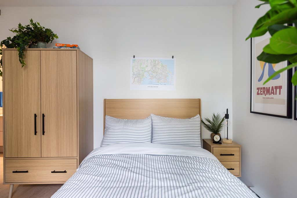 A bed with a wooden headboard and striped bedspread sits in a room with a wooden cabinet and a plant.
