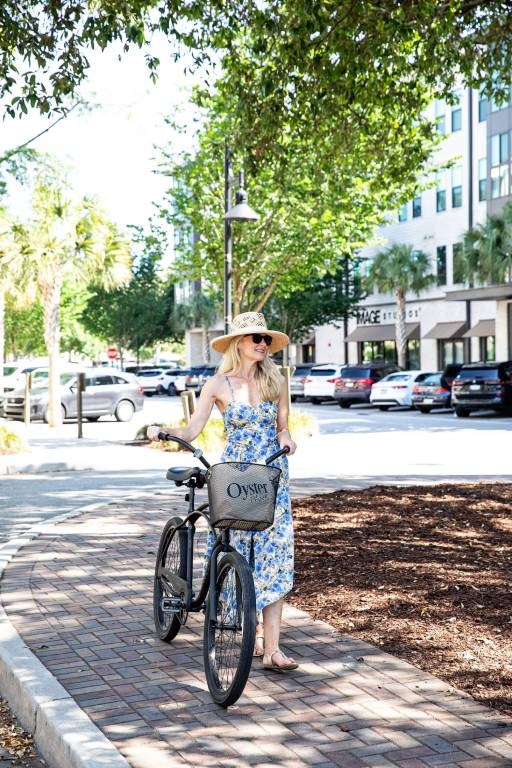 A woman in a floral dress and hat is standing next to a bicycle.