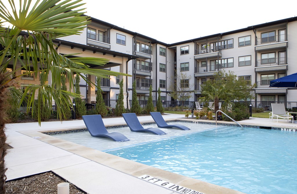 A pool surrounded by lounge chairs and trees in front of apartment buildings.