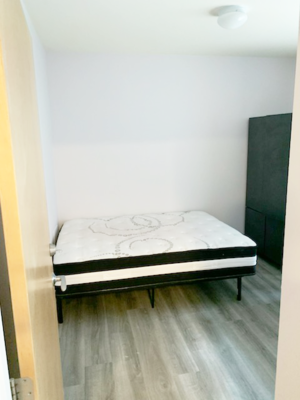 A bed with a white sheet and a black headboard is in a room with a wooden floor.