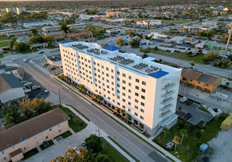 A large white building with a blue roof is surrounded by a parking lot and other buildings.