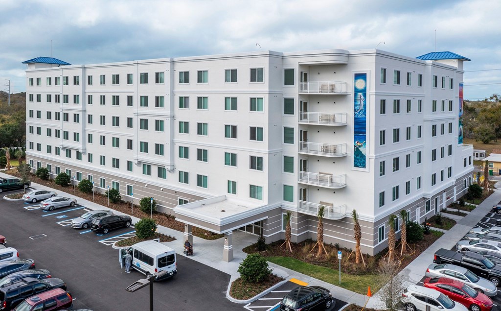 A large white hotel with a blue roof and a parking lot in front.