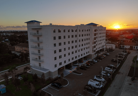 A sunset view of a white hotel with a parking lot in front.