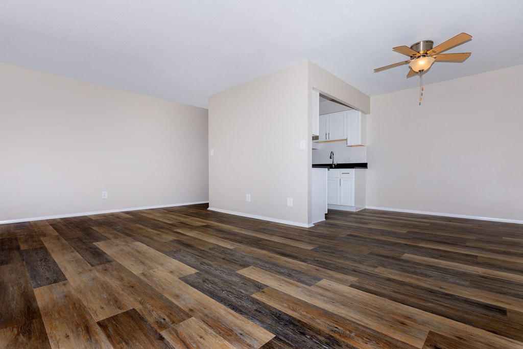 an empty living room with a ceiling fan and a kitchen in the background