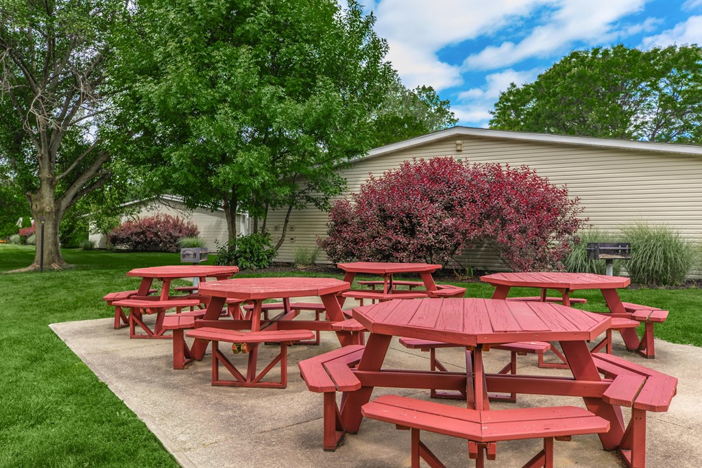 a group of red picnic tables in a grassy area with trees and a building in the