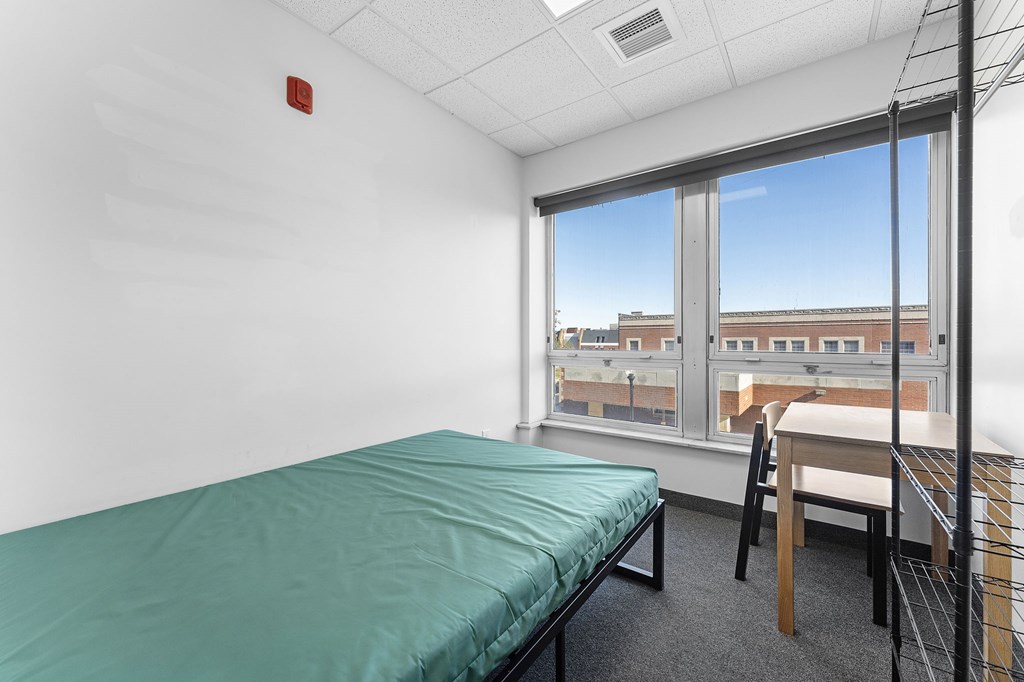 A room with a bed, desk, and chair with a window overlooking a cityscape.