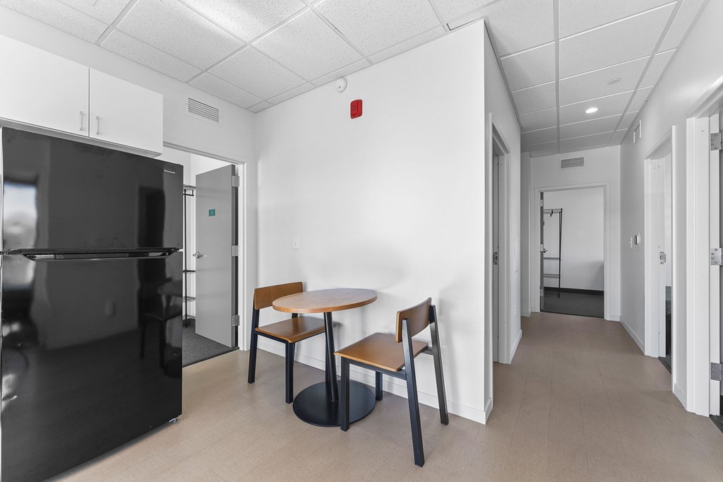 A black refrigerator is on the left in a white room with a table and two chairs.