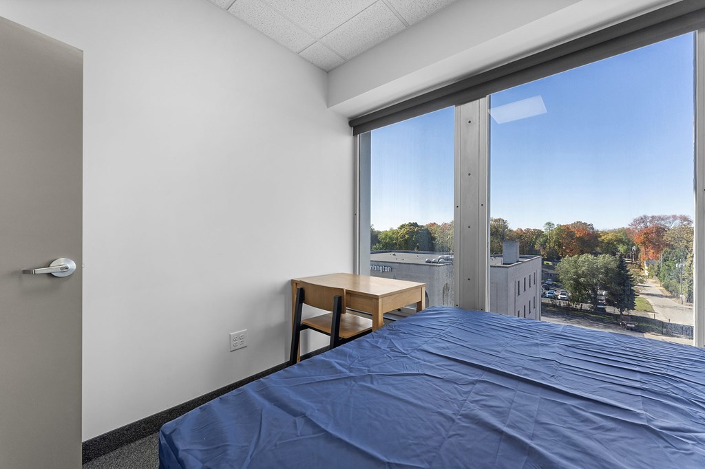 A bedroom with a bed, desk, and window overlooking a street.