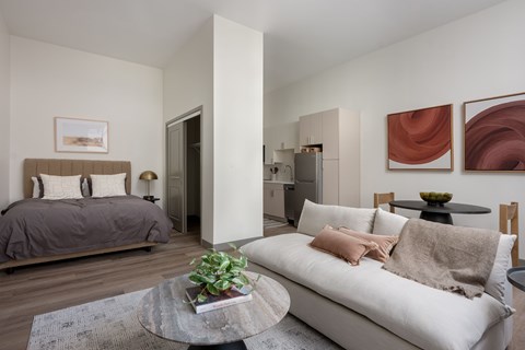 A living room with a grey couch, a bed with a grey headboard, a potted plant, and two framed artworks on the wall.