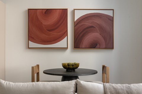 Two brown abstract paintings hang on a wall above a table with a bowl of fruit on it.