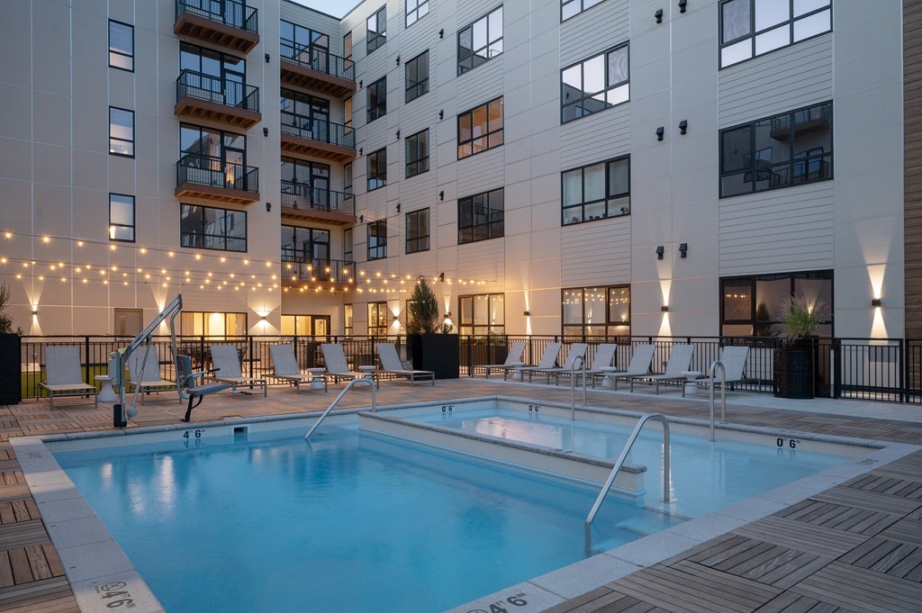 A swimming pool surrounded by lounge chairs and string lights in front of a modern apartment building.