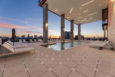 A rooftop pool area with sun loungers and a view of the city skyline at sunset.