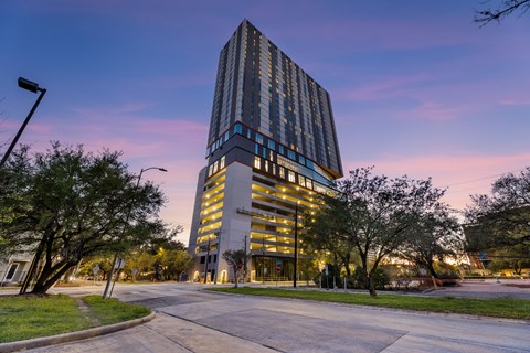 A tall building with a glass facade is illuminated from within and is surrounded by trees and a street.