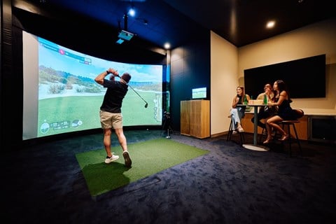 A man is playing a golf game on a screen in a room with three women watching.